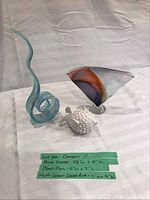 Photo showing three decorative glass and ceramic pieces on a white cloth background including a blue glass swirl sculpture, a blue fish figurine, and a multicolor triangular glass art piece, with dimension labels visible.