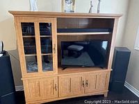 Front and angled views of light oak Sauder entertainment center with glass and solid wood doors, a TV in the center compartment, upper and lower shelves, and metal hardware.