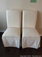 Front view of two identical ivory upholstered slipper chairs with floral fabric and gathered skirt covering the base.