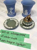 Photo showing two blue Wedgewood jasperware water cups, two green jasperware ashtrays, and green jasperware lighter with gold-tone top, all on white fabric.