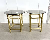 Front view of two identical brass framed end tables with round smoked glass tops, standing side by side on floor with white background.
