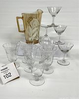 Photo showing the carnival glass jug with feather pattern alongside ten clear vintage cocktail glasses of varying styles and etched patterns.