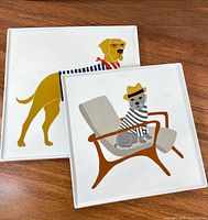 Two square ceramic platters on wooden surface showing stylized dog prints, one with a yellow dog in striped shirt and red scarf, the other with a gray dog sitting on chair.