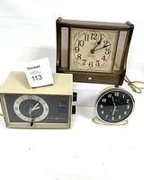 Photo of three vintage clocks: one large brown rectangular clock with mesh sides, one beige rectangular clock with a dark dial, and one small round silver alarm clock with a black dial.