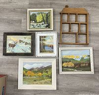 Top-down photo of five framed paintings displaying rustic primitive art landscapes and wildlife, one framed print, and a small wooden display shelf with compartments. Frames show vintage wear.