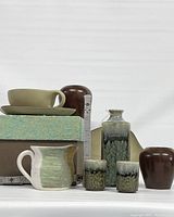 Photo showing tea cup with saucer on box, creamer jug in green and white, two green/gray speckled pottery cups, two brown pottery vases, and a ruler for scale.
