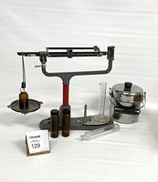View of vintage balance scale, amber glass dropper bottle on pan, three small amber bottles, clear glass measuring cylinders, glass test tube, and stainless steel tiffin carrier.