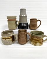 Front view of six pottery items including five mugs and one vase arranged on a flat surface with white backdrop. Various earth tones and textural finishes visible.