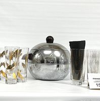 Front view of vintage chrome ice bucket with penguin design, Bakelite handles and dome lid with knob, flanked by 4 gold feather print champagne glasses on left and 4 gold starburst print champagne glasses on right.