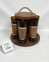 Front and side view of vintage round wooden caddy with rope handle holding six tall brown glasses with wood grip and brass bands.