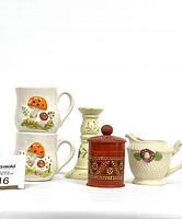 Front view of two mushroom motif mugs, wooden candle holder, red lidded wood container, and ceramic creamer