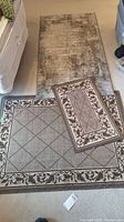 Three area rugs displayed on floor: one large mottled grey and beige rug, two smaller rugs with diamond and floral patterns.