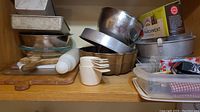 Shelf with assortment of metal bowls, cake pans, loaf pans, plastic measuring cups, wooden and plastic cutting boards, parchment bags box, rolling pin, and some plastic containers.