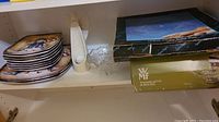 Snowman-themed ceramic platters and matching plates stacked inside a cabinet next to a clear glass bowl and white ceramic angel candle holder. Several boxes are visible nearby including a WMF box.