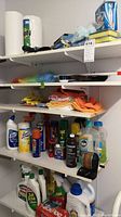 Shelf view showing multiple cleaning product bottles, cloths, and tools arranged on four white shelves.