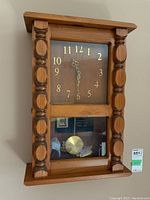 Front view of handcrafted wooden wall clock showing clock face, carved wooden columns, and visible pendulum with plaque behind glass door