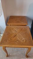 Top down view of two wooden end tables stacked slightly, showing square tops with decorative wood grain pattern and curved legs.