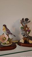 Two bird figurines on wooden stands displayed on glass shelf: one single bird with pink flower, other pair of birds on tree branch with flowers.