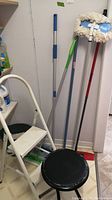 Photo showing white folding two-step stool standing next to brooms and mop with cleaning pail on floor, viewed from side angle.