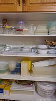 Four shelves filled with assorted plastic food storage containers, Corning Ware bowls with lids, and other kitchenware.