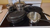 Wide shot showing all items in the set: enamel roasting pan, cast iron frying pan, four stainless steel pots (including double boiler), and splatter screen on stove.