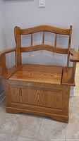 Front view of oak storage bench showing hinged seat with visible wood grain and spindle backrest