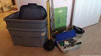 Two gray Rubbermaid Roughneck Rough Tote storage totes stacked with lids, black plunger and yellow float level, two plastic totes of hardware and household tools, and a tin tote.