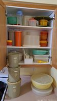 Full kitchen cabinet with stacked Tupperware bowls in various colors and sizes, clear pitcher with orange lid, yellow measuring spoon set, and smaller containers with lids.