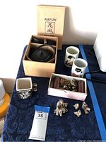 Full lot overview showing Kafuh cast iron tea set in box, two ceramic mugs, several small figurines in a box and outside, on a dark blue cloth surface.