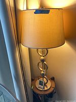 Photo showing one metal base table lamp with beige fabric drum shade on a small round wooden table next to a curtain.