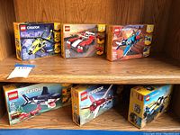 Photo showing six LEGO Creator 3-in-1 sets on wooden shelves. Boxes include helicopter, cars, planes, and a shark. All appear new and sealed.