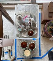 Overhead view of clear plastic bin with assorted white ceramic tea pots, pitchers, creamers, and gravy boats along with glassware and stoneware dishes below.