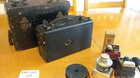 Kodak Model B movie camera, black leather carrying case, metal film reel, and various small accessories arranged on a table