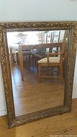 Full view of large rectangular antique mirror with gold plaster frame on wood floor showing reflection of chairs and table.