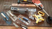 Photo showing masonry and tile tools including trowels, tile cutter, wooden level, measuring tape, hammers, and gloves arranged on carpet.