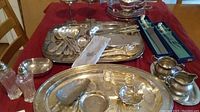 Assorted silver plate items including serving utensils on tray, salt and pepper shakers, small bowls, and glassware arranged on a red tablecloth.
