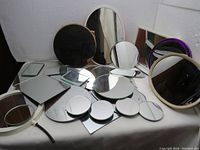 Mirrors of various shapes and sizes, including oval, round, rectangular with different colored edges, laid out on a surface.