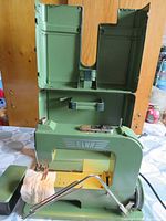 Green Elna sewing machine with attached metal base and pedal. Open green metal carrying case behind machine showing interior.