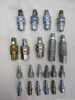 Top-down view of 23 air fittings arranged in rows; includes chrome and brass air couplers and various plug types, showing details of threading and quick connect mechanisms.