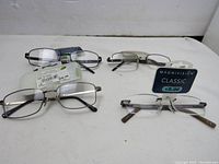Four pairs of reading glasses laid out on a white surface, two with black frames and two with rimless design. Tags indicating magnification +1.50 and +2.00 are visible.