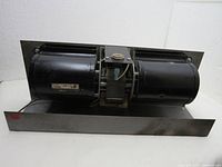 Front perspective of the squirrel cage fan showing the dual black cylindrical blower wheels attached to the motor mounted on a metal base.