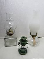 Three items: square glass oil lamp, round glass oil lamp with frosted chimney, green metal candle holder with glass chimney. All shown standing against wall.