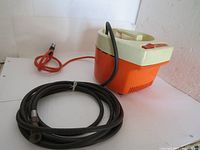 Full view of vintage Black and Decker air compressor showing orange and cream colored plastic body, attached black air hose coiled in front, and orange power cord with plug.