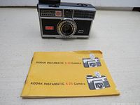 Front view of Kodak Instamatic 404 camera and yellowed instruction booklet placed side by side on flat surface.