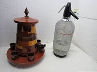 Full view of the vintage wooden liquor bottle holder with six shot glasses on a round base, and the wire mesh soda siphon bottle with chrome top and lever.