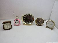 Five vintage clocks displayed side by side on white surface against a white wall; varying colors and designs visible.
