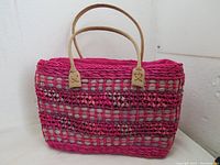 Front view of woven straw bag showing purple and pink coloring, leather handles, and open weave pattern.