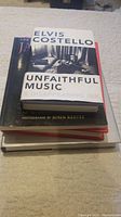 Stack of four hardcover music books, top one titled 'Unfaithful Music & Disappearing Ink' by Elvis Costello with photographs by John Reeves.