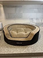 Front view of the Kirkland pet bed showing the beige cushioned interior with tufted quilting and black border, placed on a countertop.