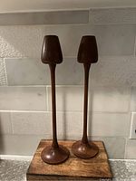 Pair of tall, slender wooden candle sticks with a round base and tapered candle holder top, set against a tiled backsplash.